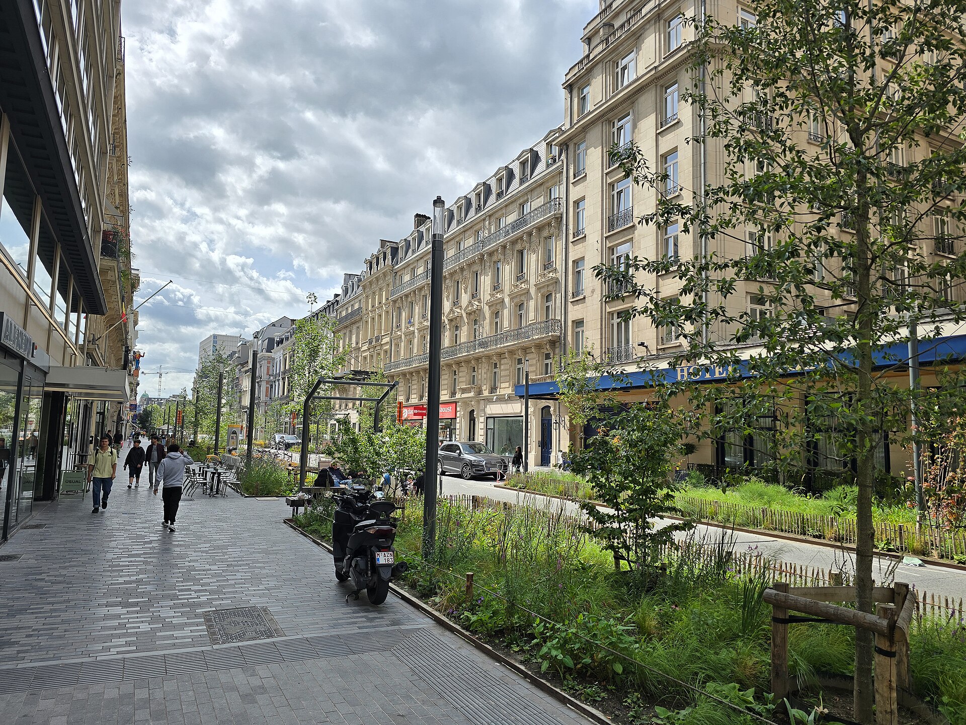 Pedestrians walk past planted beds and cafe tables along Boulevard Anspach in central Brussels, beneath rows of ornate building facades.