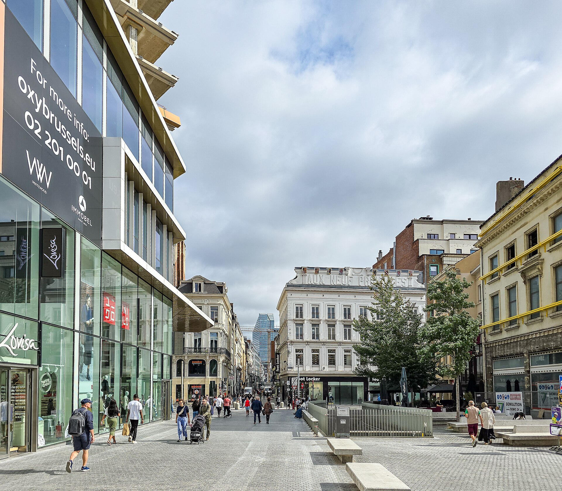 Pedestrians walk past Uniqlo and other shops on Rue Neuve in Brussels, with Aux Neuf Provinces ahead.