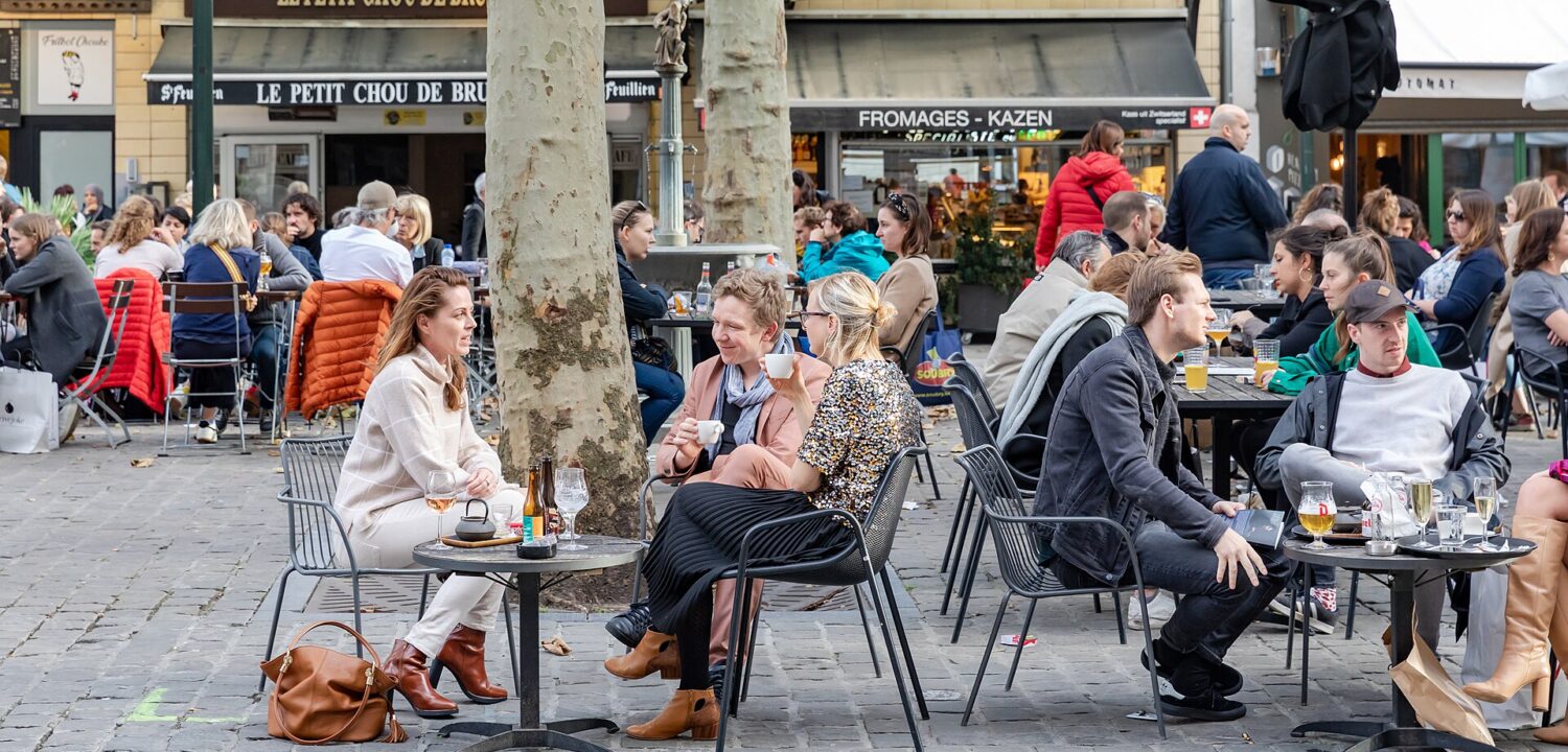 People drink coffee, wine and beer at small outdoor tables beneath trees on a busy cafe terrace in Brussels.