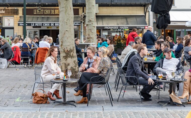 People drink coffee, wine and beer at small outdoor tables beneath trees on a busy cafe terrace in Brussels.