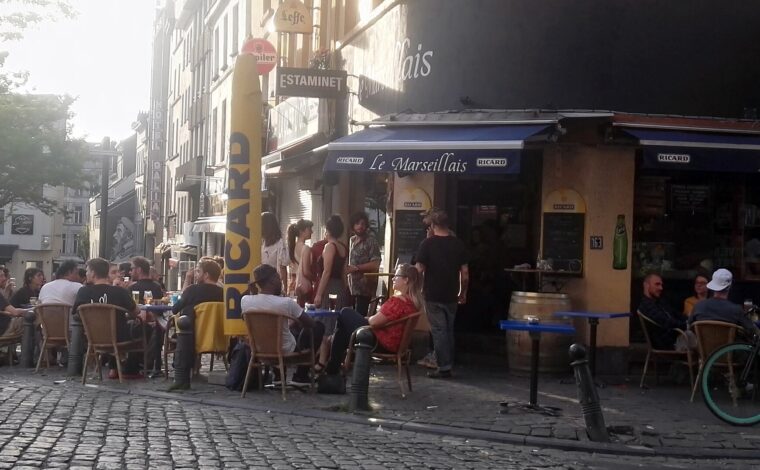 People gather around pavement tables outside Le Marseillais on Rue Blaes in Brussels, beneath blue awnings in evening sunlight.