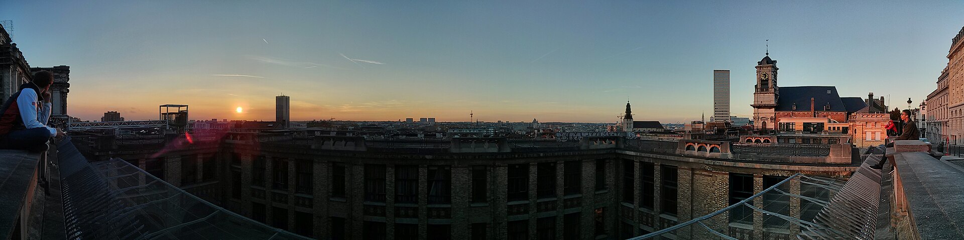People sit above a sunken terrace at Place Poelaert as the setting sun lights the wide Brussels skyline.