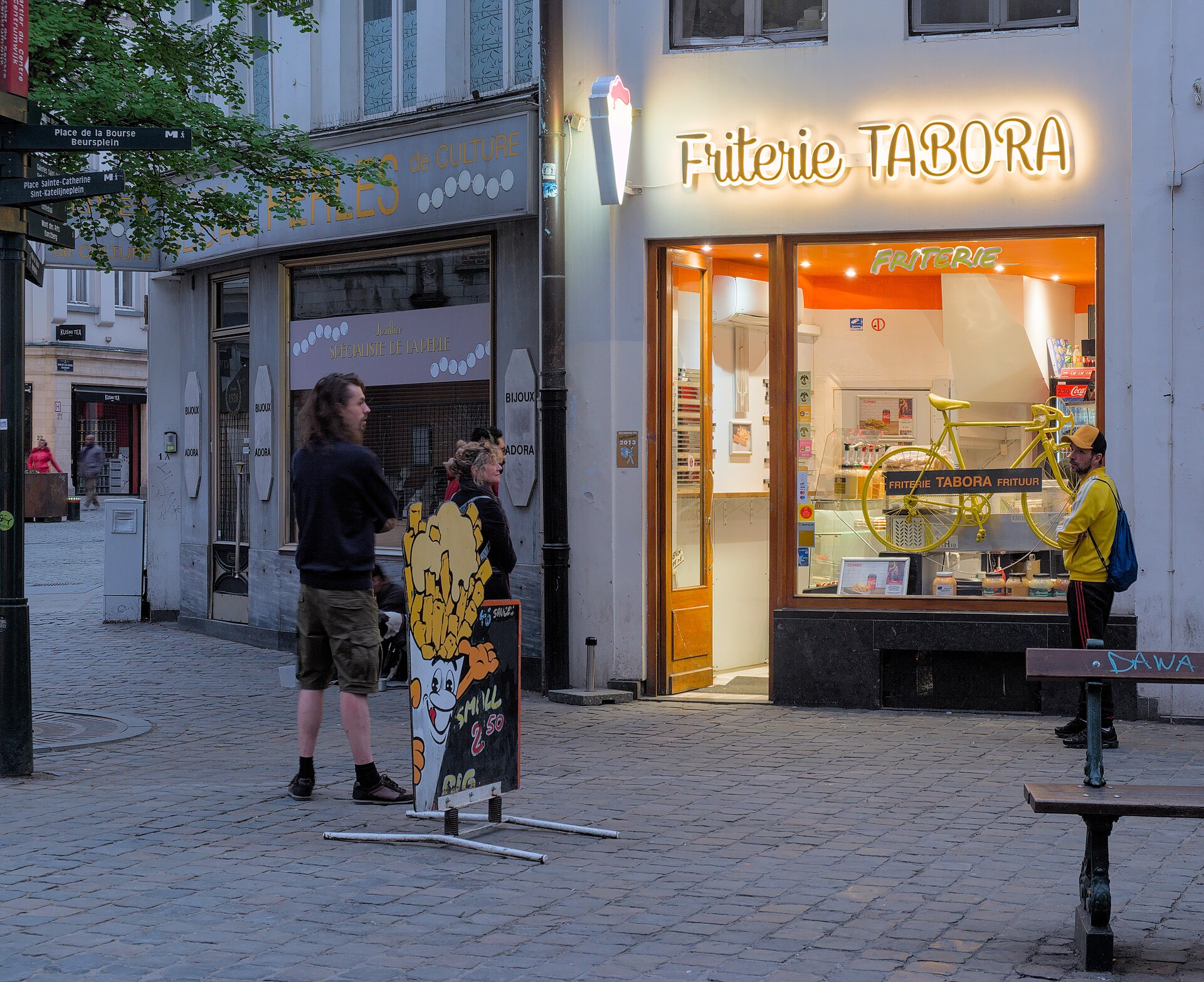 People stand outside the illuminated Friterie Tabora in Brussels, beside a fries board and an open doorway in the evening.