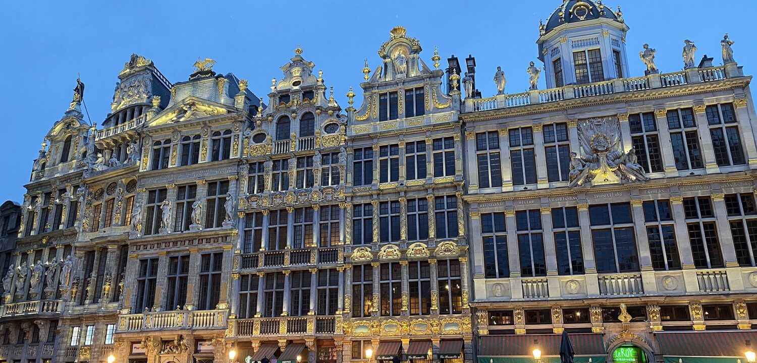 People stroll past illuminated guildhall facades and cafe terraces on the Grand-Place in Brussels at blue hour.