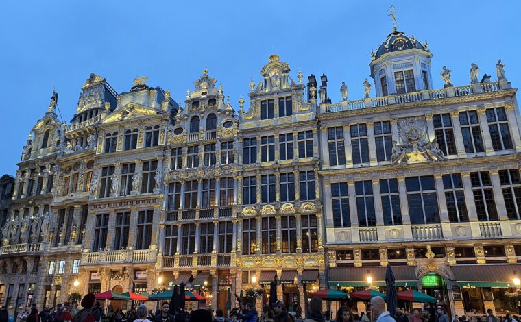 People stroll past illuminated guildhall facades and cafe terraces on the Grand-Place in Brussels at blue hour.