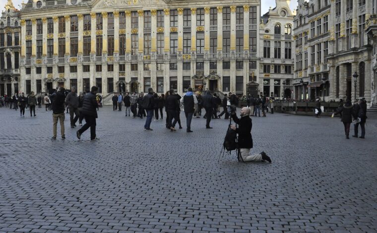 A photographer kneels among visitors on the Grand-Place in Brussels, facing the gilded facade of the House of the Dukes of Brabant.