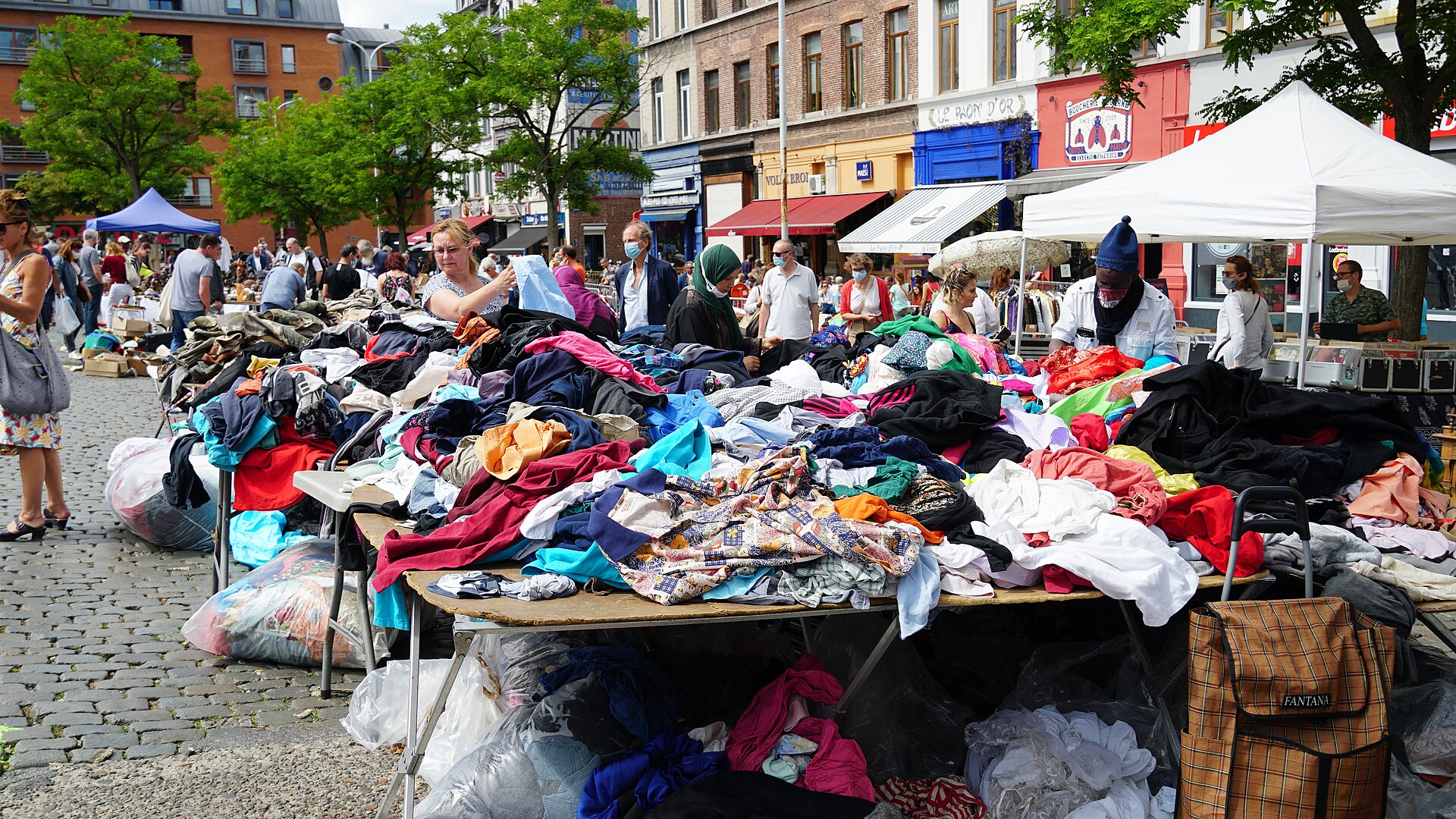 Piles of secondhand clothes cover tables at Brussels Place du Jeu de Balle while shoppers browse beside the surrounding cafes.