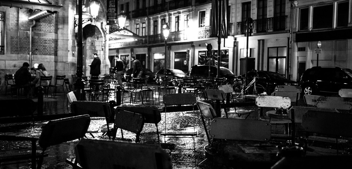 Rain glistens on outdoor tables at Zebra Bar on Place Saint-Gery in Brussels, with customers beneath illuminated street lamps.