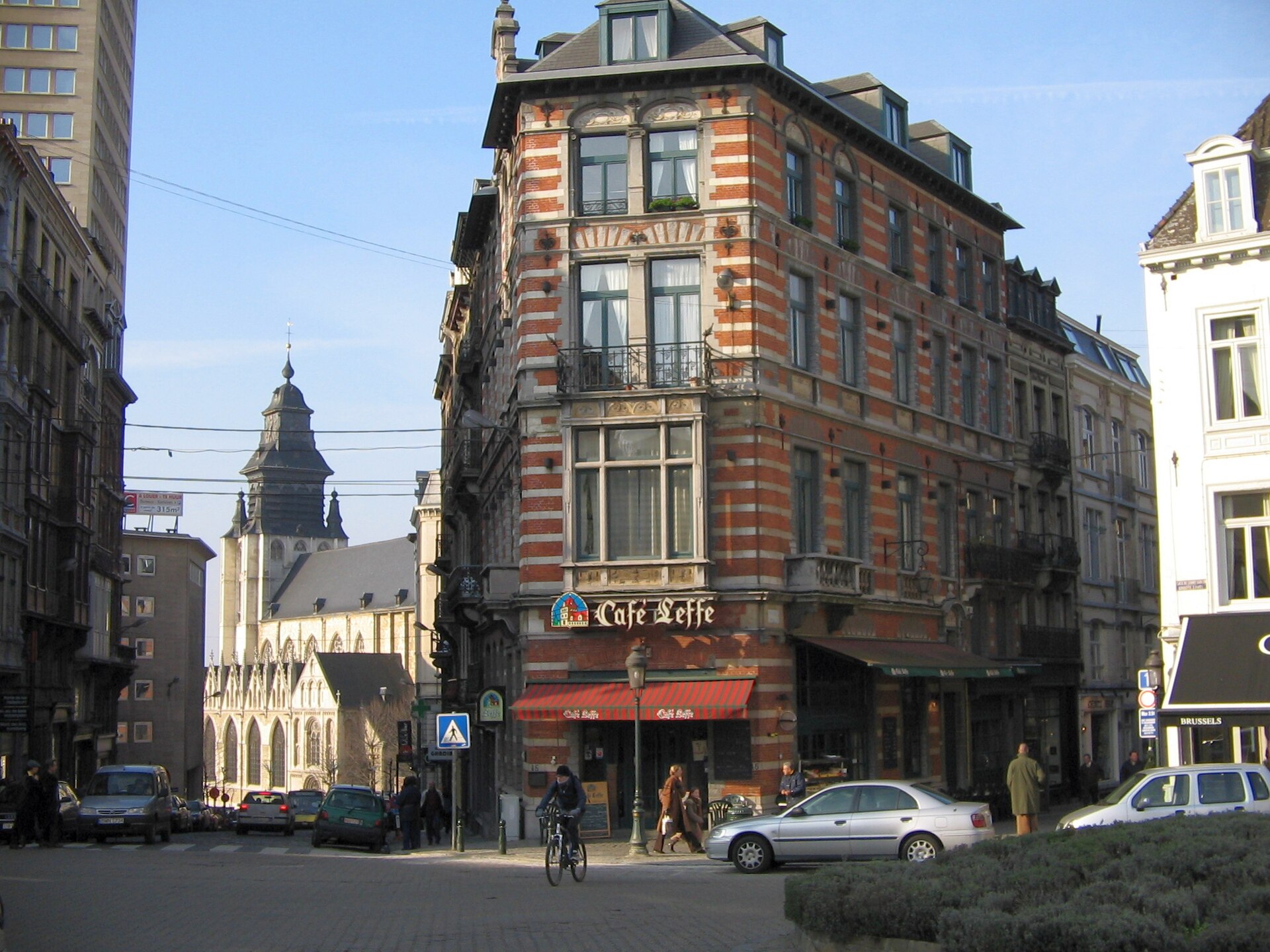 A red brick corner cafe overlooks cobblestones on Place du Grand Sablon in Brussels, with a church beyond.