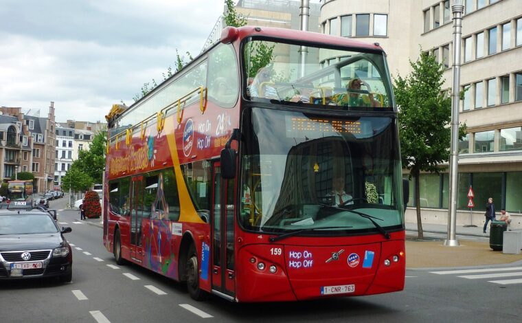 A red City Sightseeing Brussels open-top bus passes city buildings, with passengers upstairs and a taxi beside it.