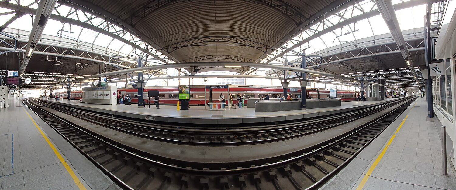 A red international train stands beside passengers at Brussels-Midi, beneath a broad steel canopy spanning the platforms and railway tracks.