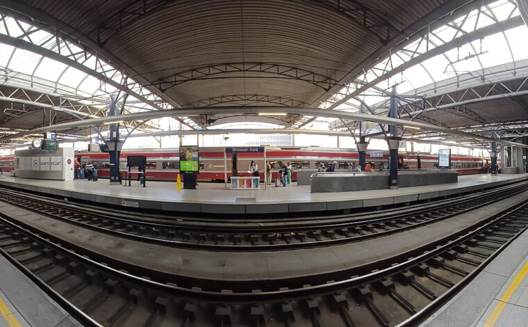 A red international train stands beside passengers at Brussels-Midi, beneath a broad steel canopy spanning the platforms and railway tracks.
