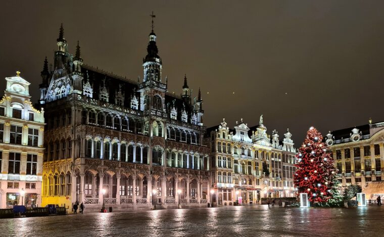 A red-lit Christmas tree stands beside illuminated guildhouses and Brussels City Museum on the Grand-Place, reflected in wet cobblestones.