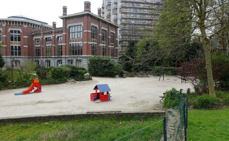 A red playhouse, slide and swings stand in a sandy playground beside historic brick buildings in Brussels’ Leopold Park.