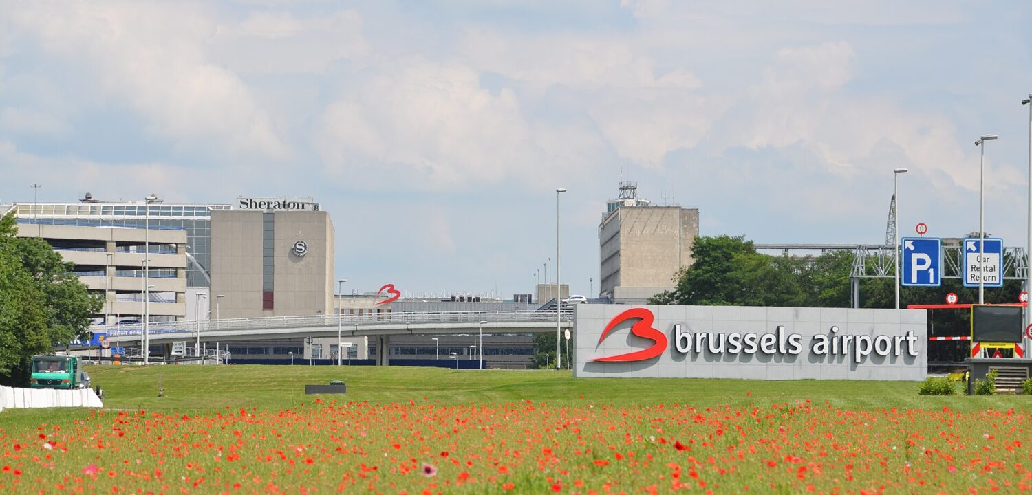 Red poppies frame the Brussels Airport sign, terminal approach roads and Sheraton hotel at Zaventem beneath a pale sky.