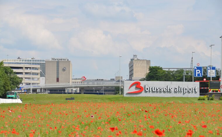 Red poppies frame the Brussels Airport sign, terminal approach roads and Sheraton hotel at Zaventem beneath a pale sky.