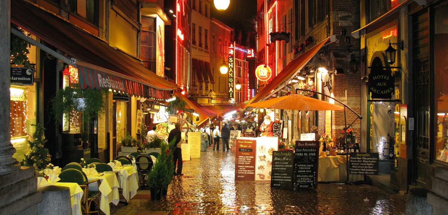 Restaurant lights reflect on rain-wet cobblestones along Rue des Bouchers in Brussels, where pedestrians pass tables beneath striped awnings.