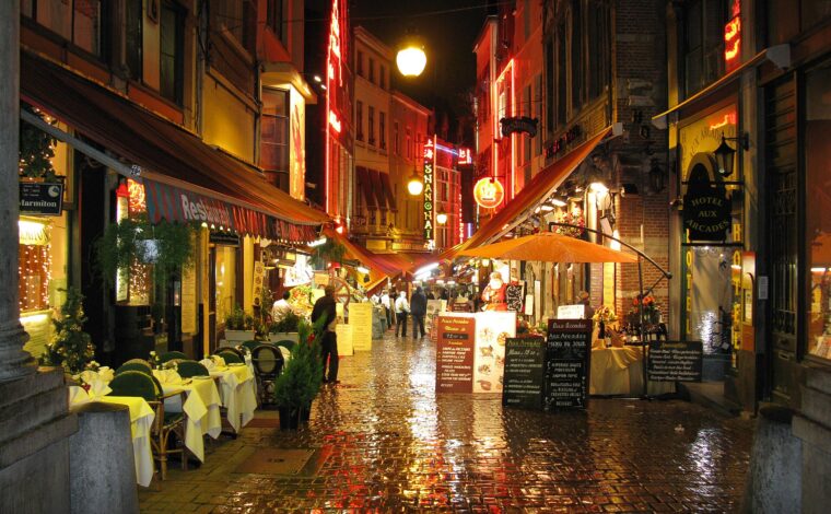 Restaurant lights reflect on rain-wet cobblestones along Rue des Bouchers in Brussels, where pedestrians pass tables beneath striped awnings.