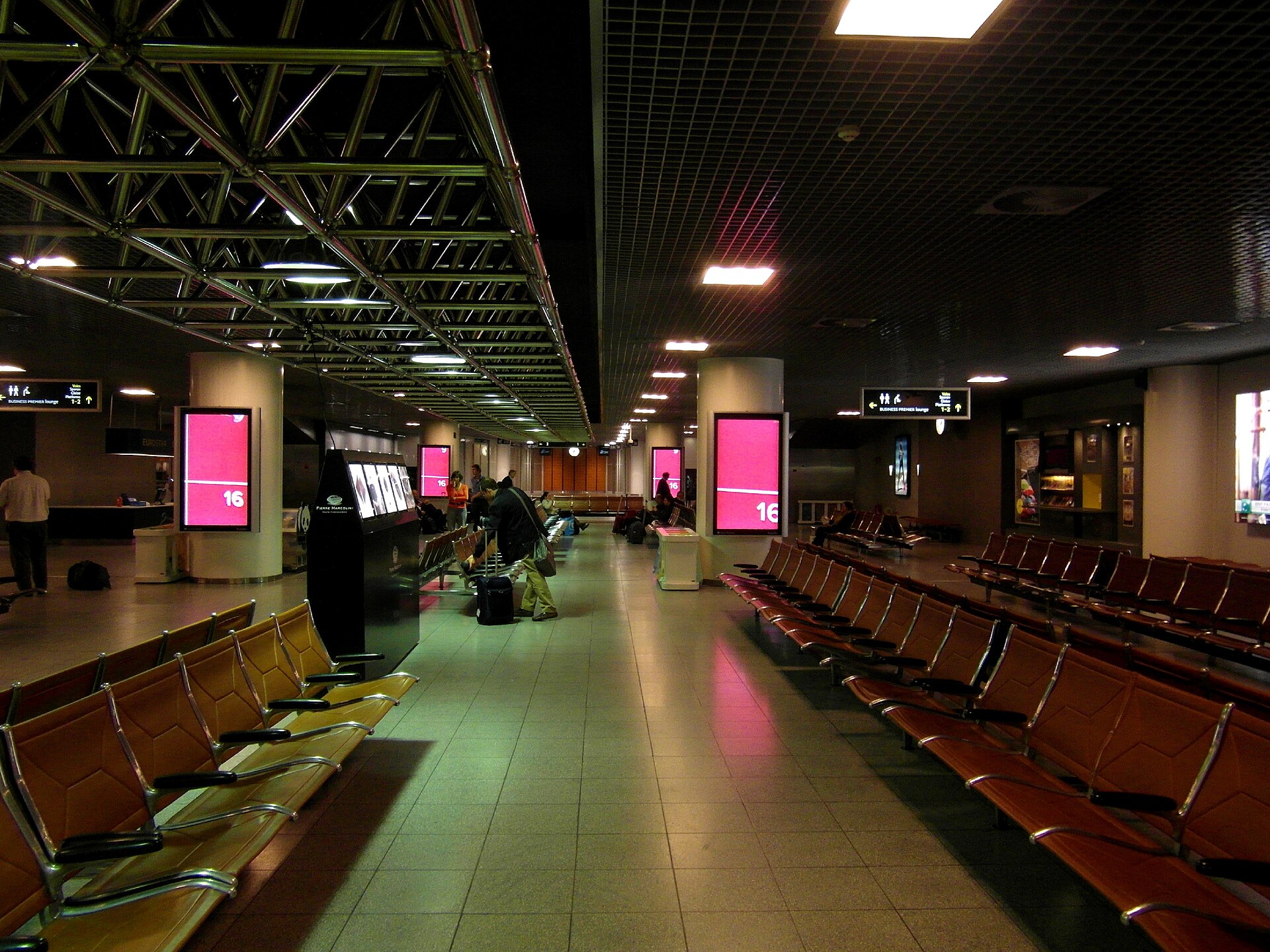 Rows of brown seats fill the Eurostar waiting hall at Brussels-Midi, with travellers and illuminated panels beneath ceiling girders.