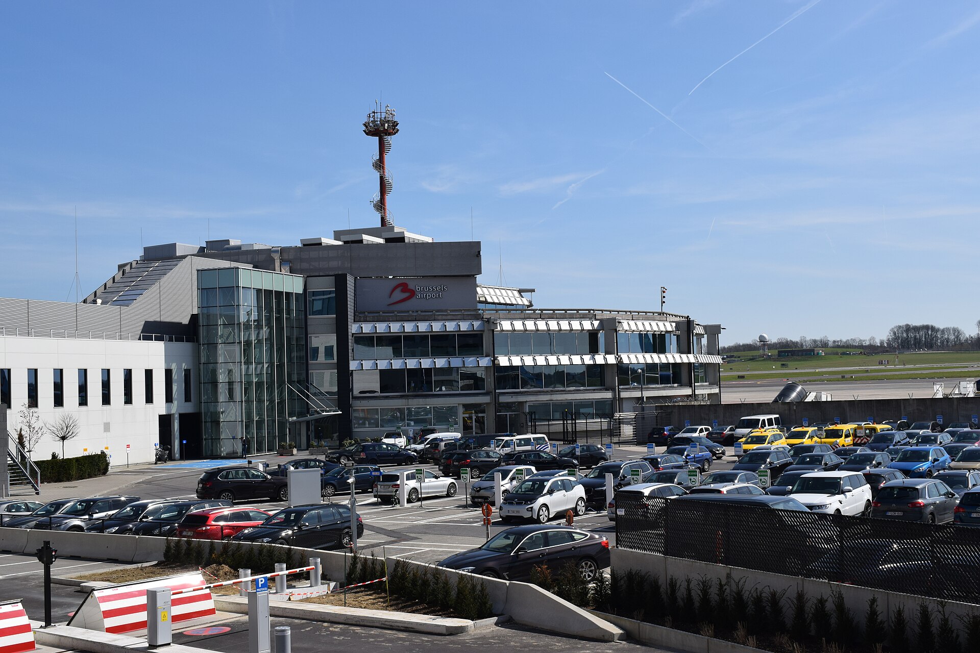 Rows of parked cars fill a barrier-controlled car park beside a Brussels Airport building, with the airfield visible beyond.