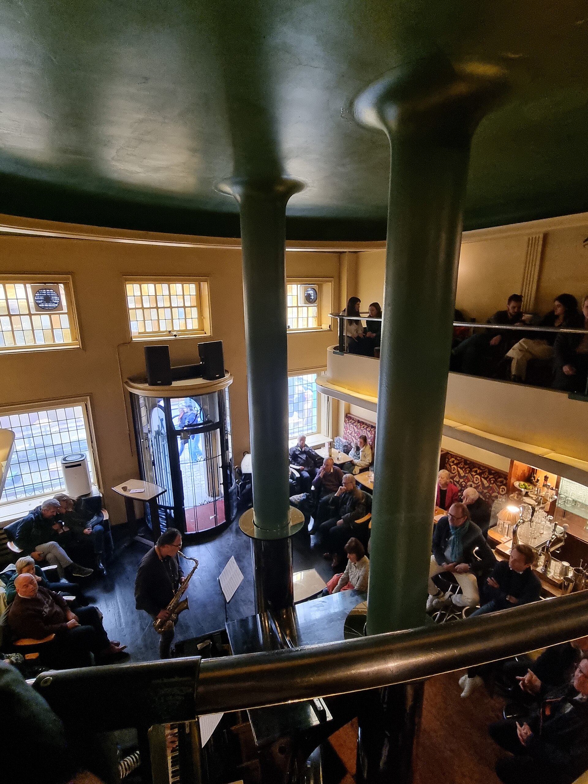 A saxophonist plays below green columns and curved balconies inside L'Archiduc in Brussels, with listeners seated around the piano.