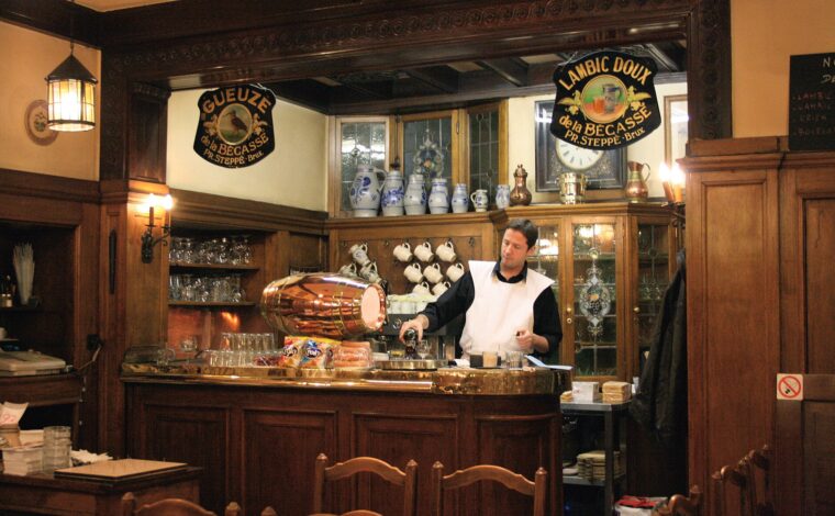 A server pours a drink behind the wooden bar at Brussels A la Becasse, beneath gueuze and lambic signs.