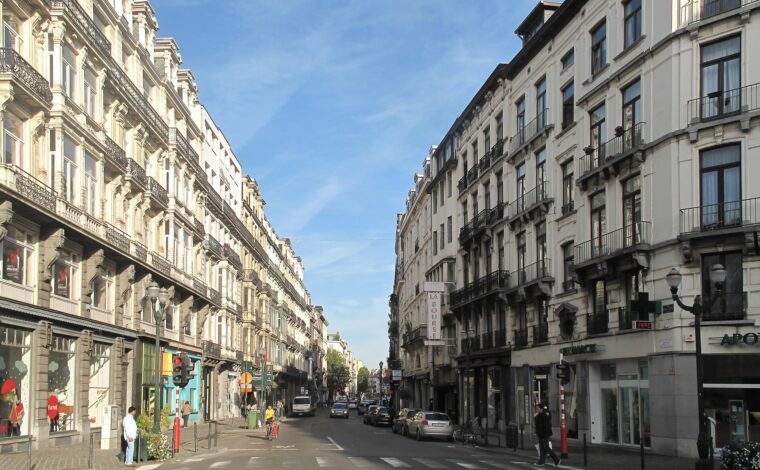 Shop windows and ornate balconies line Rue Antoine Dansaert in Brussels as pedestrians and a cyclist pass.