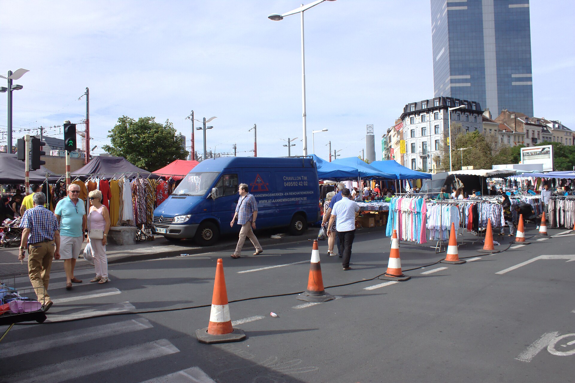 Shoppers browse clothing stalls at Brussels Midi Market beneath blue canopies, with the South Tower rising behind nearby buildings.