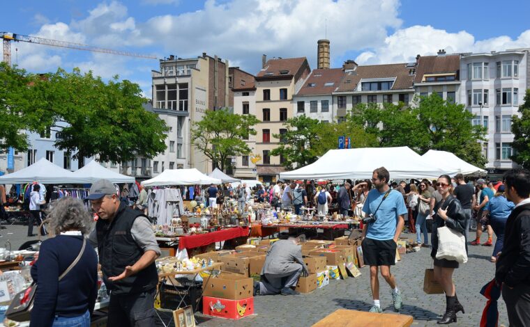 Shoppers browse ornaments and clothing at Place du Jeu de Balle in Brussels, with white tents and the public baths behind.