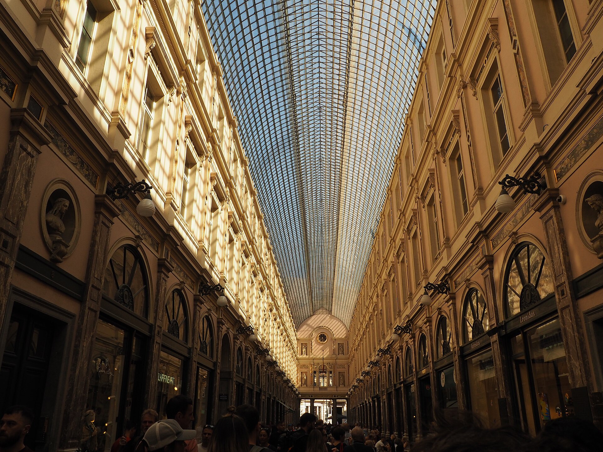 Shoppers fill Brussels' Galeries Royales Saint-Hubert beneath a long glass vault, cream pilasters and rows of arched shopfronts.