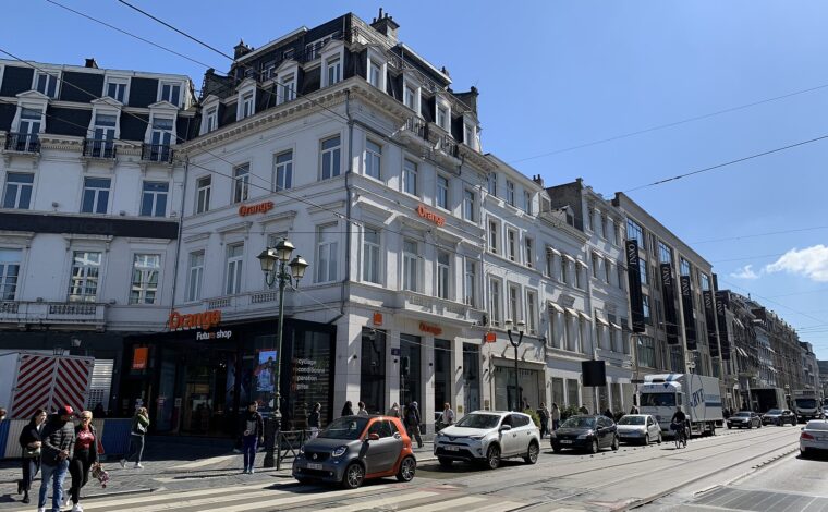 Shoppers pass white facades, Orange and Inno storefronts beside tram rails at the entrance to Avenue Louise in Brussels.