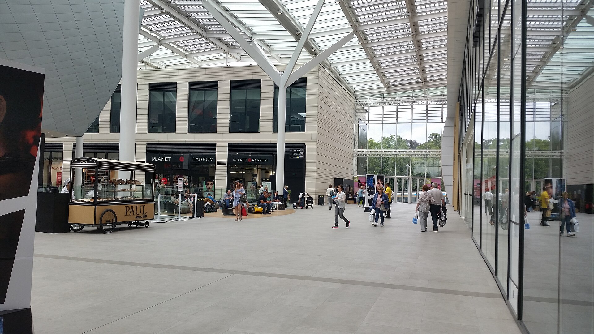 Shoppers walk beneath the glass roof at Docks Bruxsel in Brussels, passing a bakery kiosk and bright storefronts.