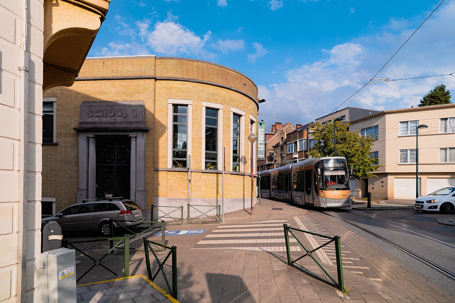 A silver and gold STIB tram rounds a corner beside School 1 and residential buildings in Evere, Brussels.