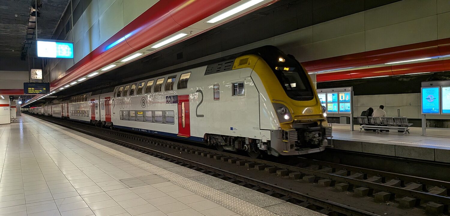 A silver SNCB double-deck train with red doors stands beside the underground platform at Brussels Airport Zaventem station.
