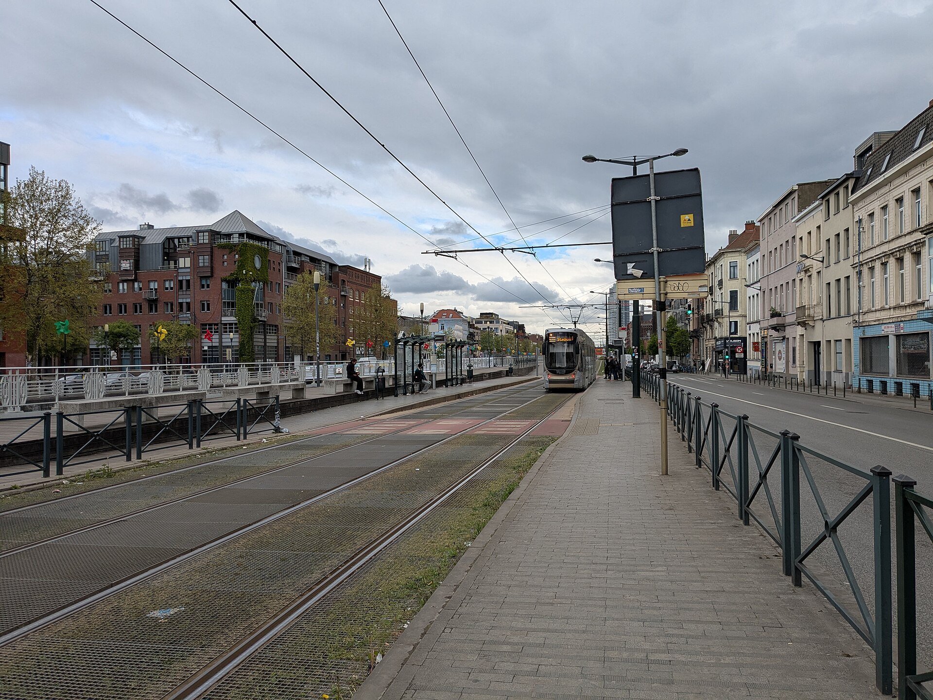 A silver STIB tram approaches Porte de Flandre in Brussels beside platforms, overhead wires and rows of buildings.