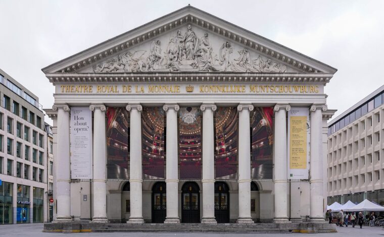 Six columns frame opera banners beneath the sculpted pediment of La Monnaie opera house on its Brussels square.