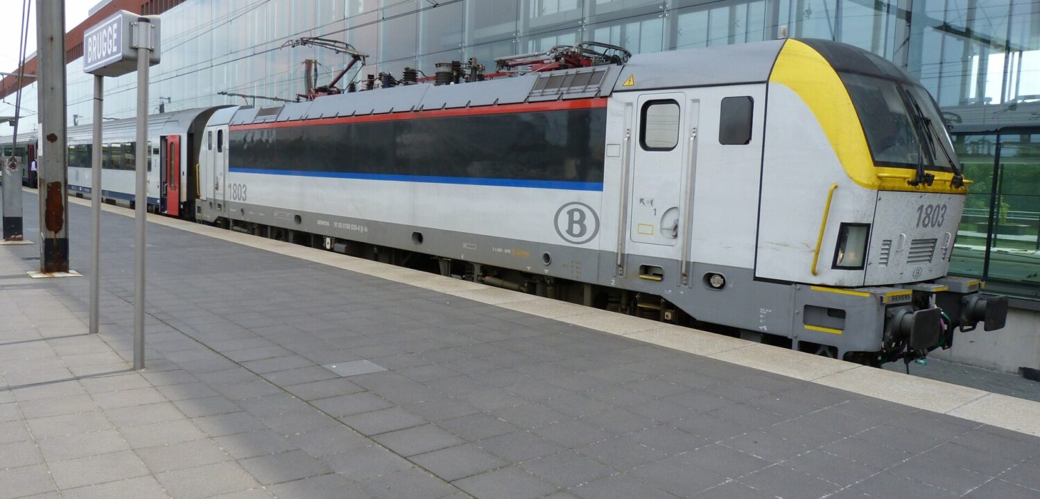 SNCB locomotive 1803 and passenger coaches stand beside a Brugge station sign and the glass-fronted railway station building.