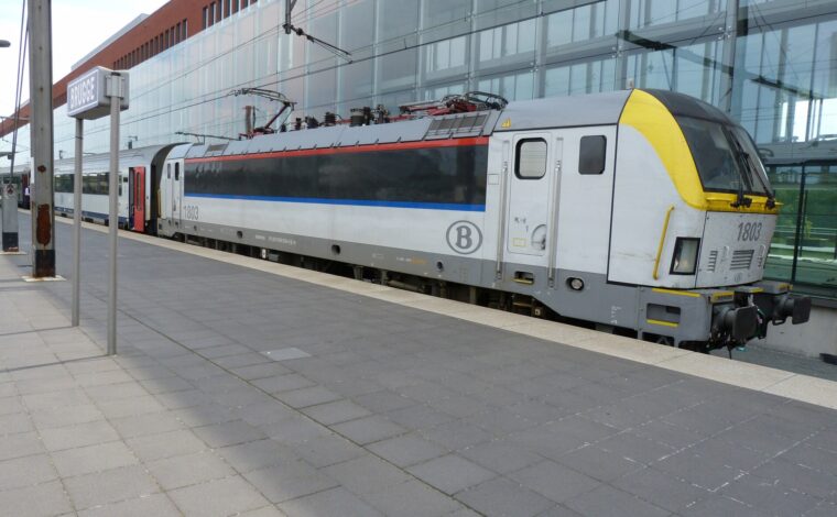 SNCB locomotive 1803 and passenger coaches stand beside a Brugge station sign and the glass-fronted railway station building.