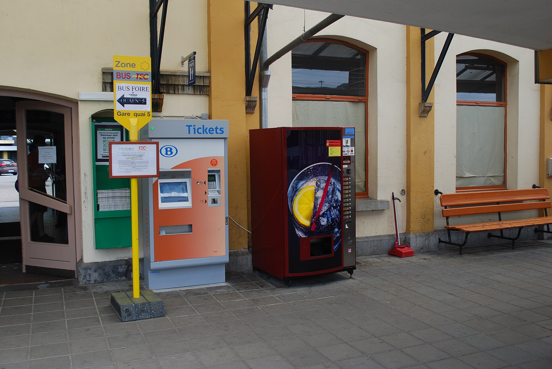 An SNCB ticket machine stands beside a drinks vending machine and an orange bench at Libramont station in Belgium.