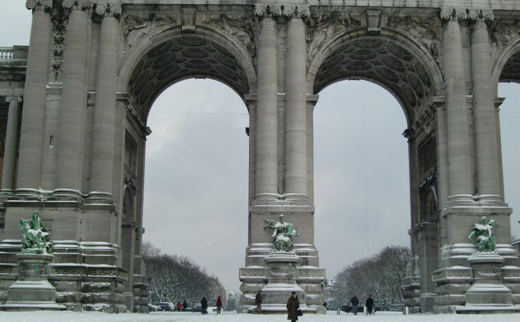 Snow blankets the ground beneath Brussels' Cinquantenaire arch, where bundled visitors walk past bronze sculptures and massive stone columns.