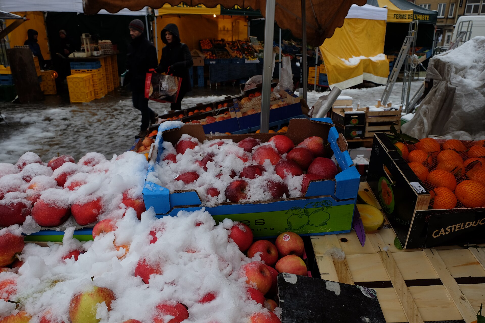 Snow-dusted crates of red apples and oranges on a market stall in Brussels, with shoppers carrying bags past more produce stalls and yellow tents behind