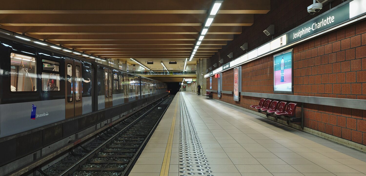 A STIB metro train stands beside the signed Joséphine-Charlotte platform in Brussels, with red seats along the brick wall.