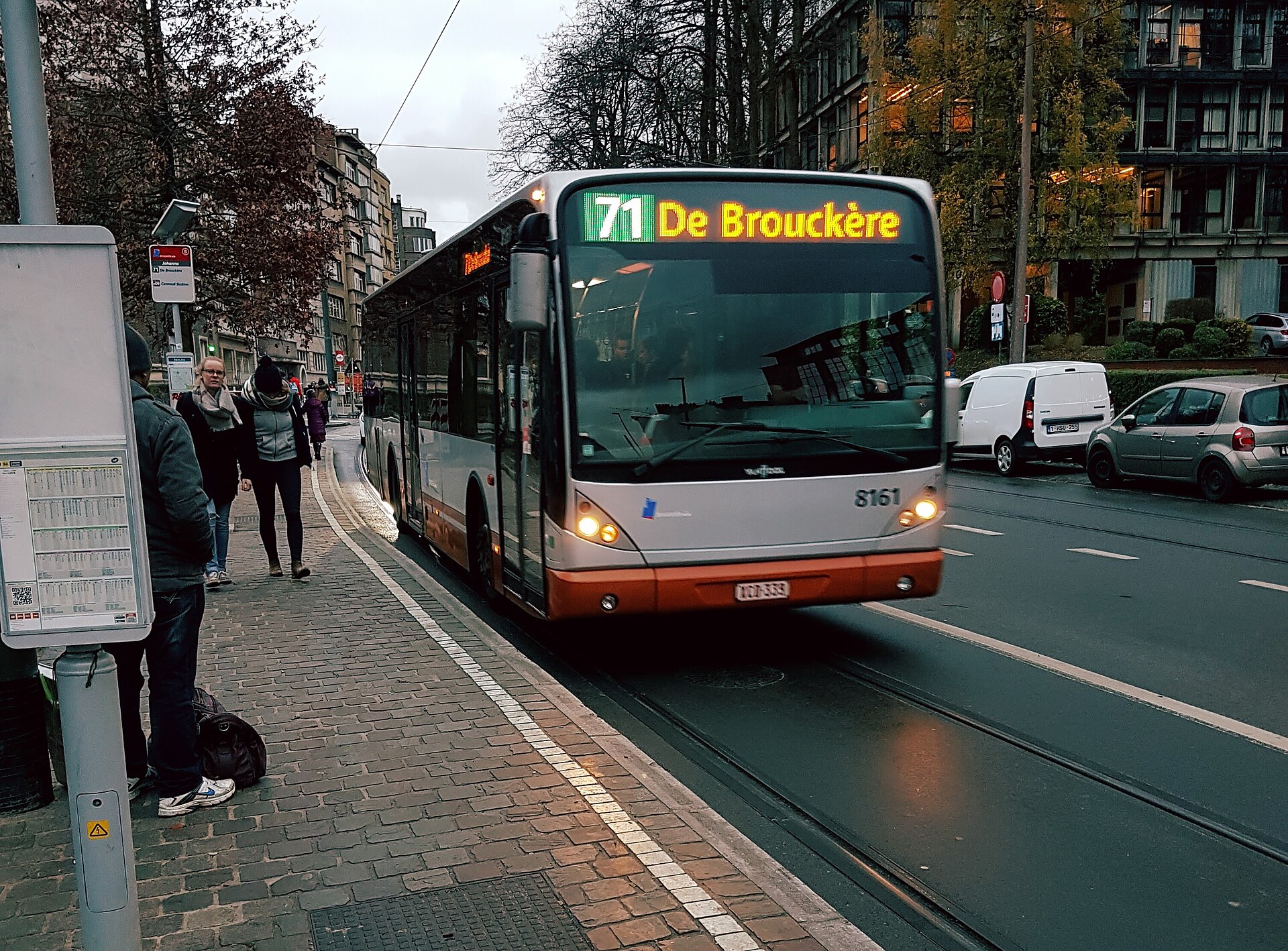 A STIB route 71 bus to De Brouckère approaches a stop on a tree-lined street in Ixelles, Brussels.