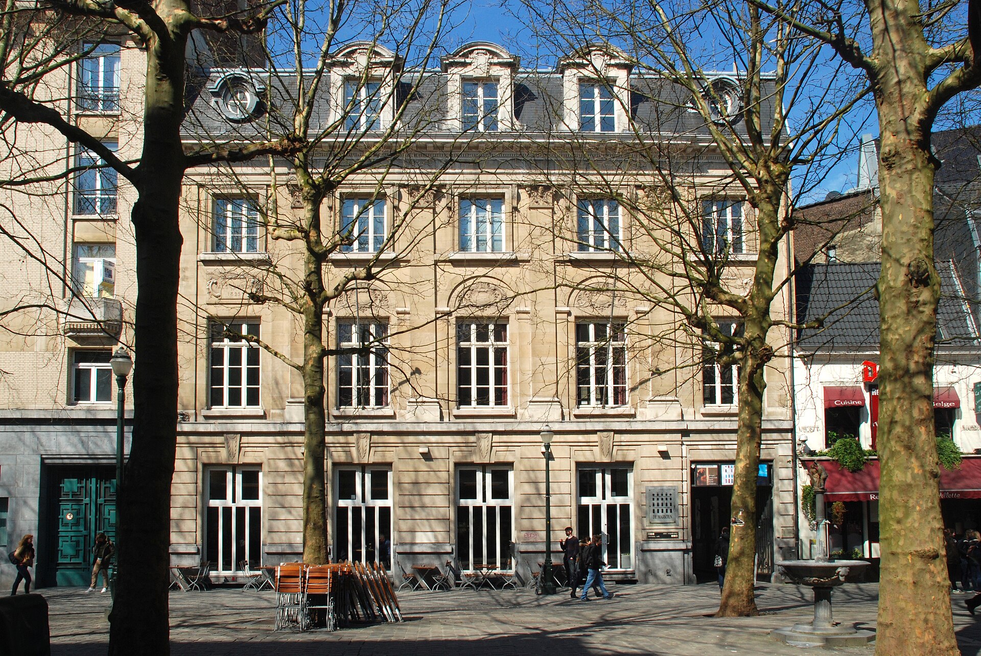The stone facade of the De Markten cultural centre behind bare plane trees on the Place du Vieux Marché aux Grains in central Brussels