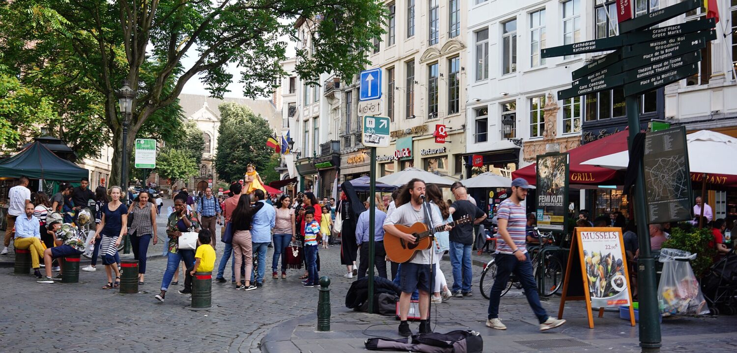 A street musician plays guitar on Rue du Marché aux Herbes in Brussels as pedestrians pass historic facades and cafe terraces.