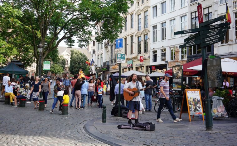 A street musician plays guitar on Rue du Marché aux Herbes in Brussels as pedestrians pass historic facades and cafe terraces.