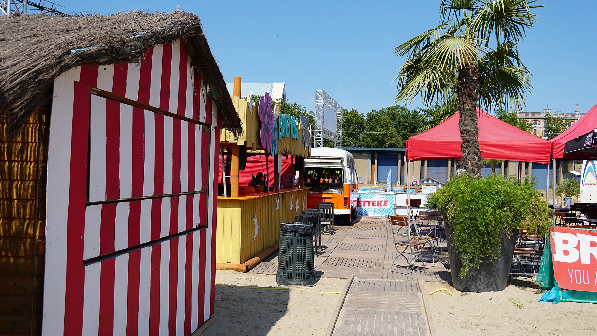 Striped thatched beach hut, bar stall and palm tree on a sandy boardwalk at the Bruxelles-les-Bains summer beach in Brussels on a sunny day.