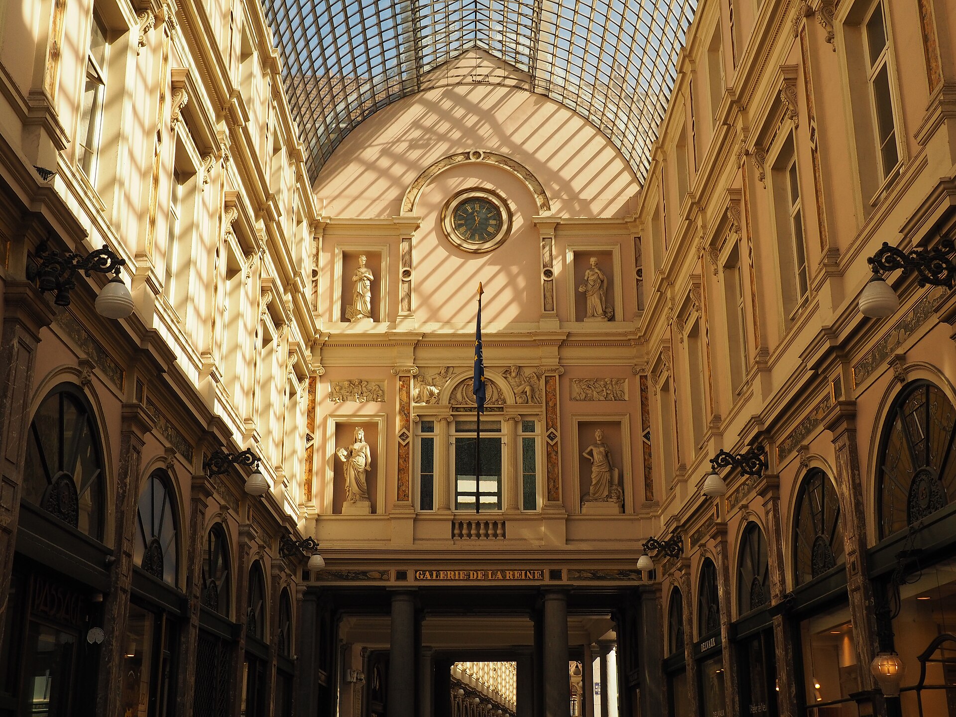 Sunlight crosses the glass roof above the clock, statues and Galerie de la Reine sign in Brussels' Saint-Hubert galleries.