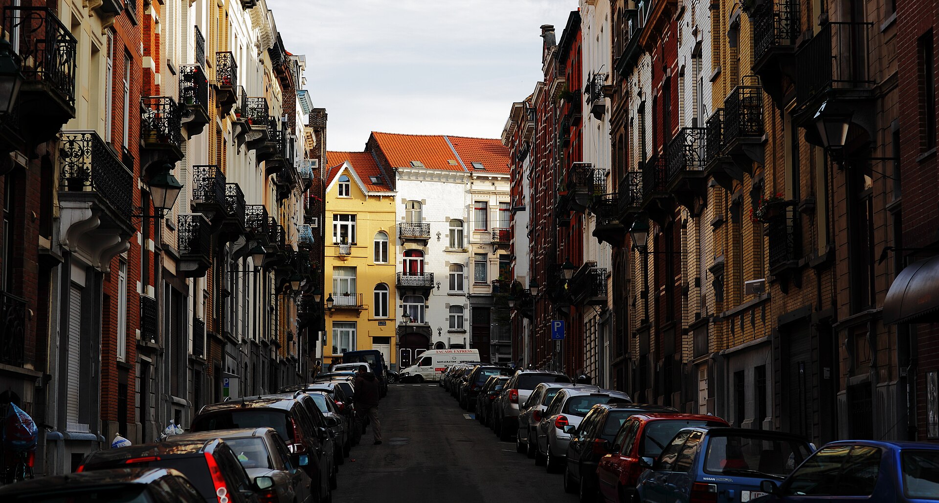 Townhouses with iron balconies line a quiet residential street in Saint-Gilles, Brussels, with parked cars along both kerbs.