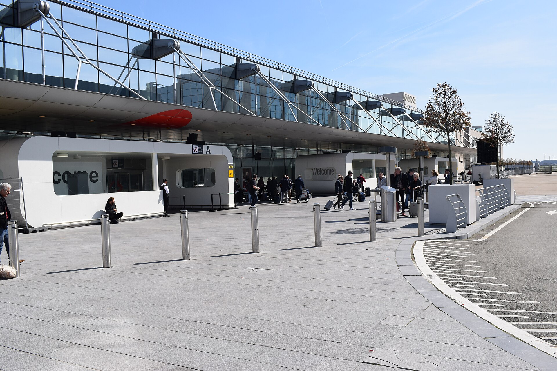 Travellers with wheeled luggage gather outside the glass-fronted departures entrance at Brussels Airport on a sunny day.